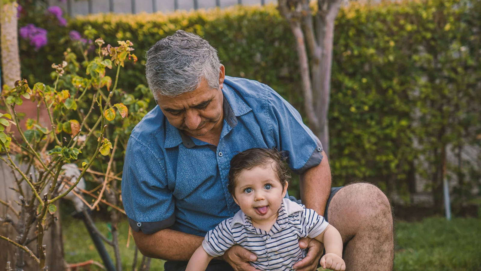 An older man holding a smiling baby in a garden.
