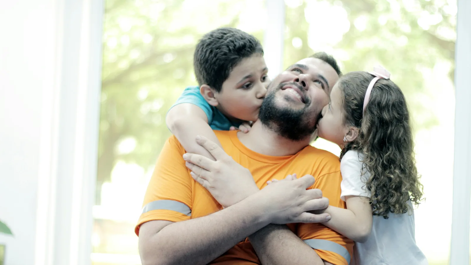 A smiling man as a boy and a girl hug him and kiss his cheeks.